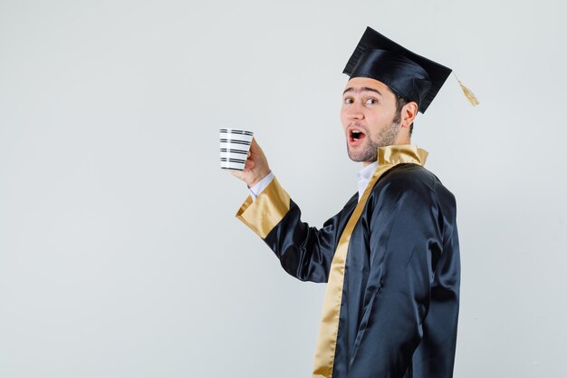 Jovem segurando um copo de bebida em uniforme de pós-graduação e parecendo alegre.