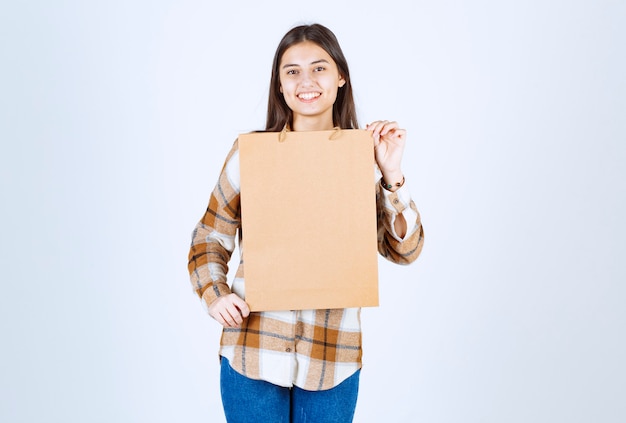 Jovem, segurando um pacote de papel artesanal e em pé sobre uma parede branca.