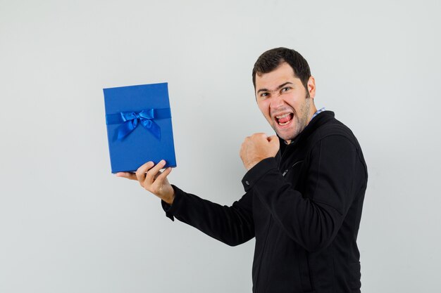 Jovem segurando uma caixa de presente, mostrando o gesto do vencedor na camisa, jaqueta e parecendo feliz