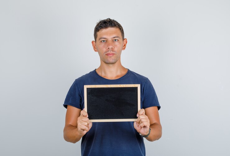 Foto grátis jovem segurando uma pequena lousa em camiseta azul escuro e olhando sério. vista frontal.