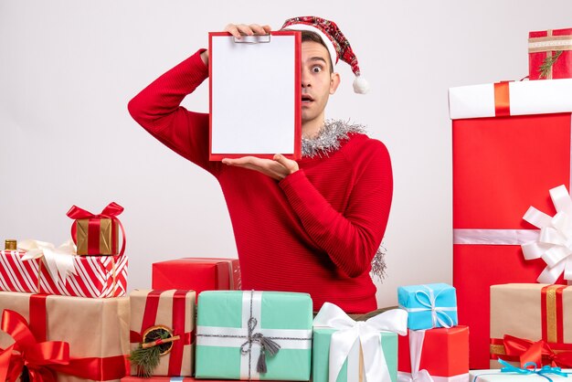 Jovem segurando uma prancheta sentado em frente aos presentes de natal