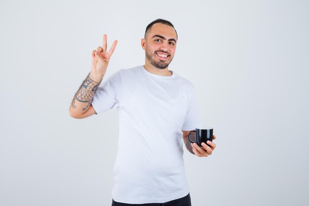 Jovem segurando uma xícara de chá e mostrando o símbolo da paz em uma camiseta branca e calça preta e parecendo feliz