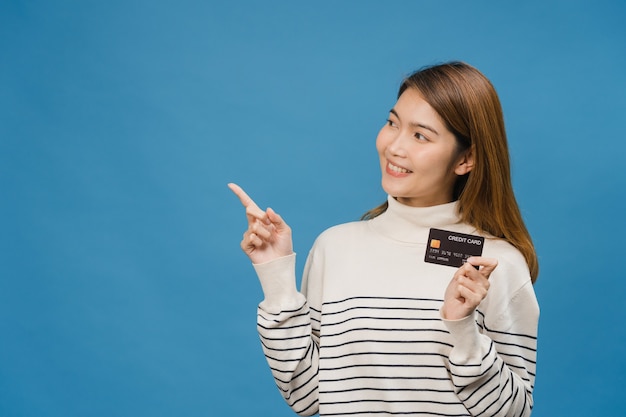 Jovem senhora asiática mostra cartão de crédito com expressão positiva, sorri amplamente, vestida com roupas casuais sentindo felicidade e estande isolado na parede azul