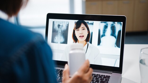 Jovem senhora asiática usando computador laptop fala sobre uma doença em videoconferência com consulta on-line do médico sênior na sala de estar em casa.