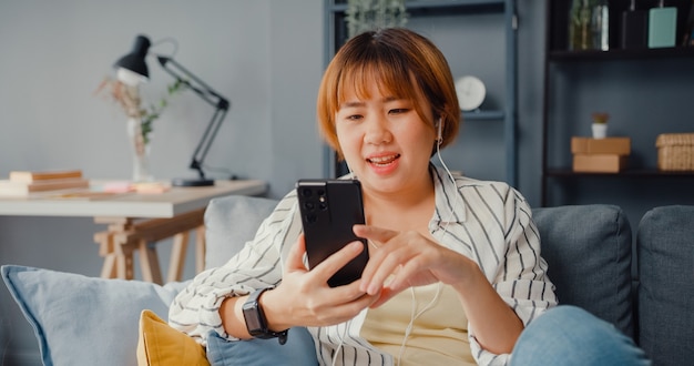 Jovem senhora asiática usando telefone inteligente para falar por videochamada com a família no sofá na sala de estar em casa
