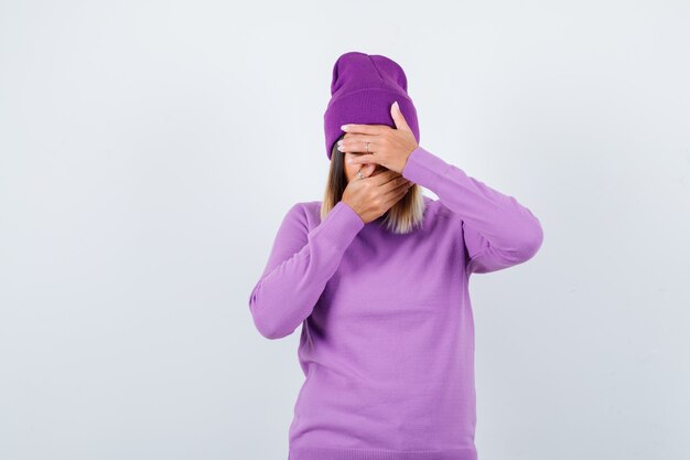 Jovem senhora com as mãos no rosto de suéter roxo, gorro e parecendo triste. vista frontal.