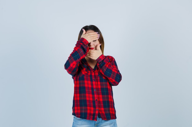 Jovem senhora com as mãos no rosto em camisa xadrez e olhando triste, vista frontal.