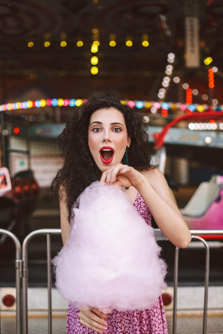 Foto grátis jovem senhora com cabelo escuro encaracolado em vestido de pé com algodão doce rosa na mão e olhando espantado para a câmera no parque de diversões