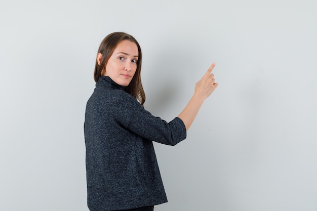 Jovem senhora com camisa apontando para cima enquanto olha para a frente e parece sensata, vista traseira