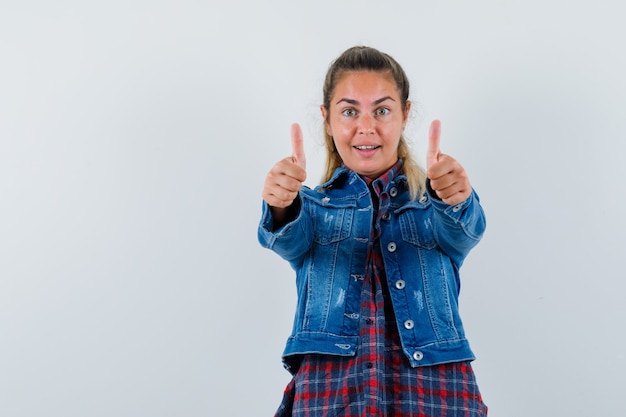 Jovem senhora com camisa, jaqueta mostrando dois polegares para cima e parecendo feliz, vista frontal.