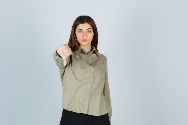 Jovem senhora com camisa, saia mostrando o polegar para baixo e parecendo decepcionada, vista frontal.