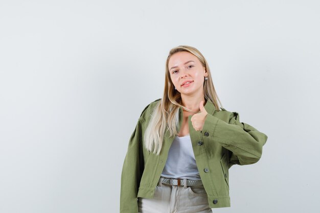 Jovem senhora com casaco, camiseta mostrando o polegar e olhando alegre, vista frontal.