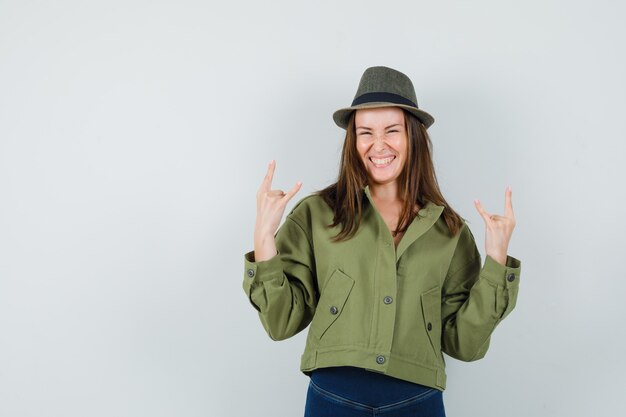 Jovem senhora com chapéu de calça jaqueta fazendo o símbolo do rock e parecendo feliz