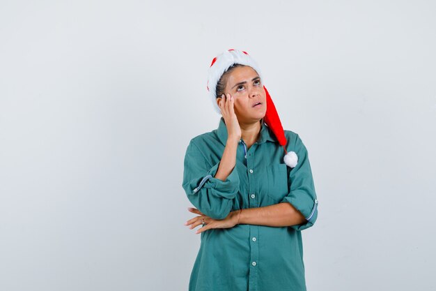 Jovem senhora com chapéu de Natal, camisa com a mão perto do rosto e olhando pensativa, vista frontal.