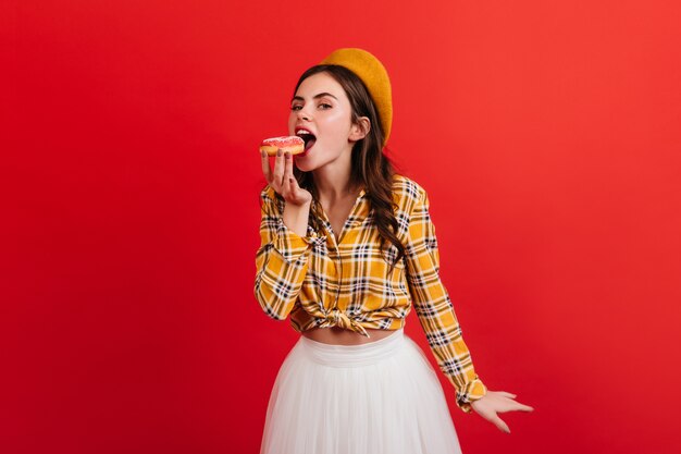 Jovem senhora com saia branca fofa morde donut. Parisiense na boina e blusa amarela, posando na parede vermelha.