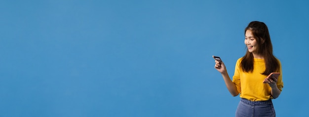 Foto grátis jovem senhora da ásia usando telefone e cartão de crédito com expressão positiva, sorria amplamente, vestida com roupas casuais e ficar isolado sobre fundo azul. fundo de banner panorâmico com espaço de cópia.