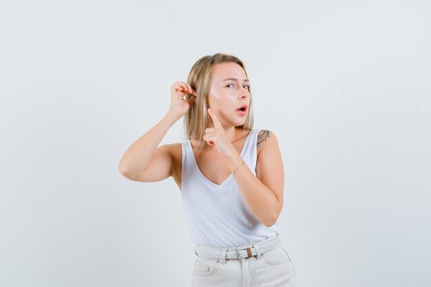 Jovem senhora de blusa branca apontando para a orelha e parecendo concentrada