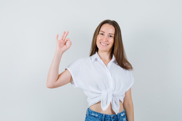 Jovem senhora de blusa branca, mostrando o gesto de ok e olhando feliz, vista frontal.