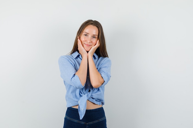 Jovem senhora de camisa azul, calça segurando as bochechas nas palmas das mãos e linda