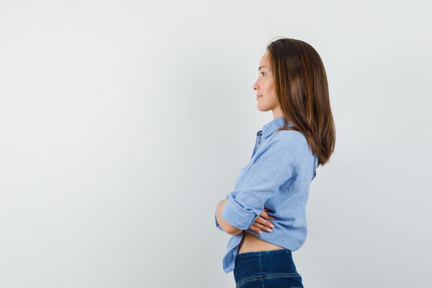 Jovem senhora de camisa azul, calças em pé com os braços cruzados e parecendo esperançosa