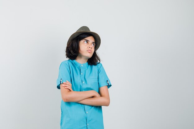 Jovem senhora de camisa azul, chapéu em pé com os braços cruzados olhando para o lado e parecendo complicado