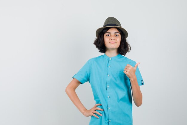 Jovem senhora de camisa azul, chapéu mostrando o polegar e parecendo feliz