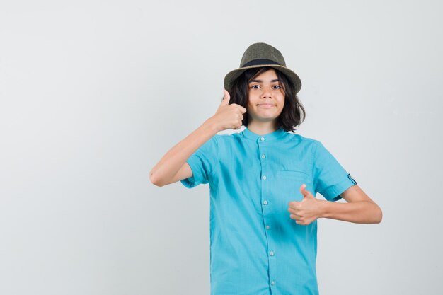 Jovem senhora de camisa azul, chapéu mostrando o polegar e parecendo feliz