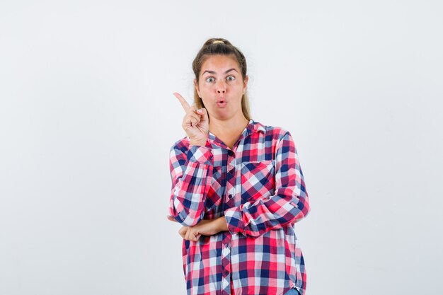 Jovem senhora de camisa quadrada, apontando para cima e parecendo surpresa, vista frontal.