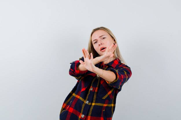 Jovem senhora de camisa quadrada, mostrando o gesto de pare e olhando perplexa, vista frontal.