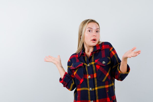 Jovem senhora de camisa quadrada, mostrando um gesto impotente e parecendo perplexa, vista frontal.