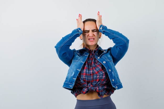 Jovem senhora de camisa quadriculada, jaqueta jeans, sofrendo de dor de cabeça e parecendo dolorida, vista frontal.