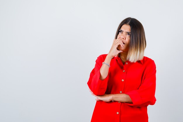 Jovem senhora de camisa vermelha enorme roendo as unhas e parecendo pensativa, vista frontal.