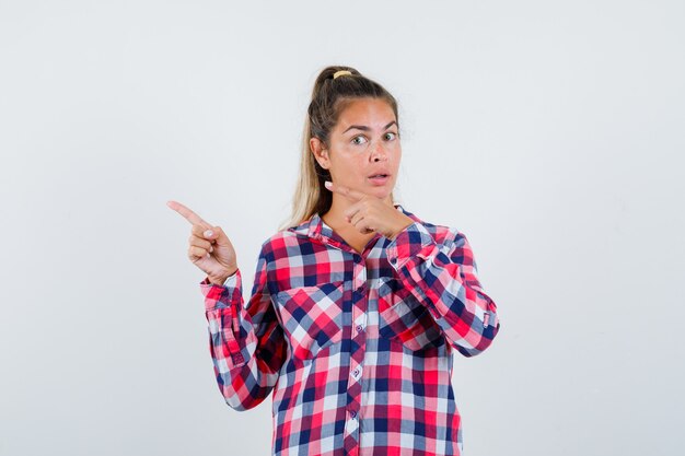 Jovem senhora de camisa xadrez, apontando para o canto superior esquerdo e parecendo hesitante, vista frontal.