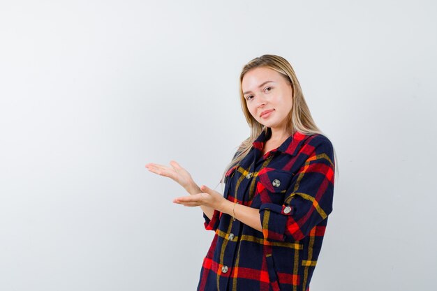 Jovem senhora de camisa xadrez, dando boas-vindas a algo e parecendo confiante, vista frontal.