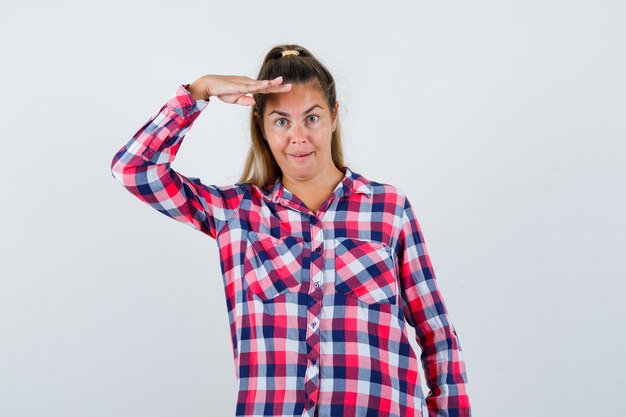 Jovem senhora de camisa xadrez, olhando para a câmera com a mão na cabeça e olhando espantado, vista frontal.