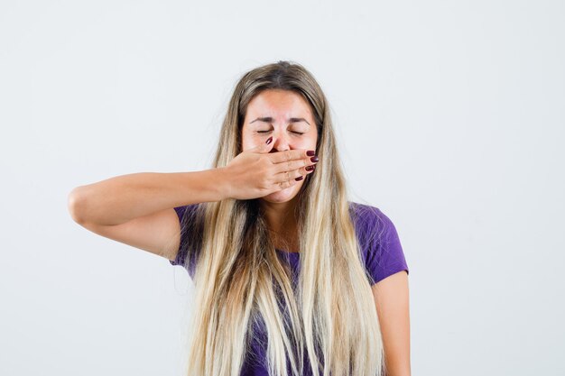 Jovem senhora de camiseta violeta, segurando a mão na boca e parecendo triste, vista frontal.