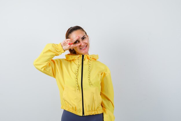 Jovem senhora de casaco amarelo, mostrando o sinal de vitória no olho e olhando alegre, vista frontal.