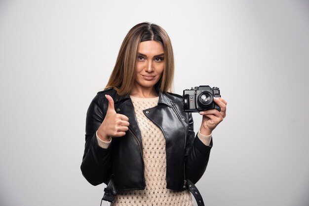 Jovem senhora de jaqueta de couro preta tirando fotos com a câmera de forma positiva e sorridente.