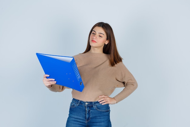 Jovem senhora de suéter bege, jeans segurando uma pasta azul e olhando com foco, vista frontal.