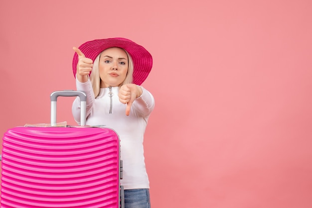 Jovem senhora em pé atrás de uma mala rosa fazendo sinais de positivo e negativo