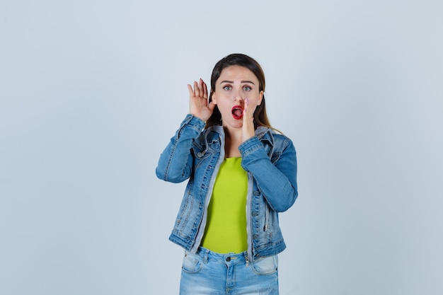 Jovem senhora em roupa de jeans, mantendo a mão perto da boca enquanto ouve o segredo e parece encantada, vista frontal.