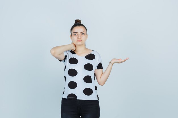 Jovem senhora em t-shirt, jeans fingindo mostrar algo, segurando a mão no pescoço e parecendo confiante, vista frontal.