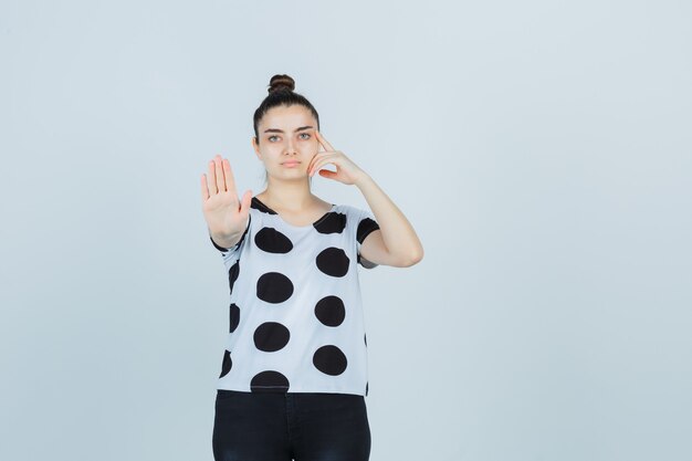 Jovem senhora em t-shirt, jeans, mostrando o gesto de parar e olhando séria, vista frontal.