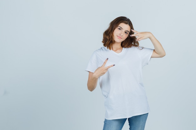 Jovem senhora em t-shirt, jeans, mostrando sinal de vitória e olhando alegre, vista frontal.