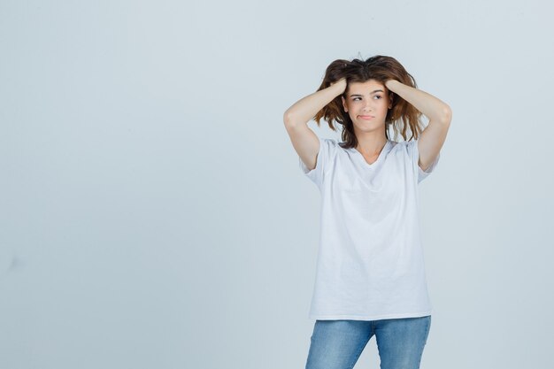 Jovem senhora em t-shirt, jeans, segurando as mãos na cabeça e olhando esquecido, vista frontal.