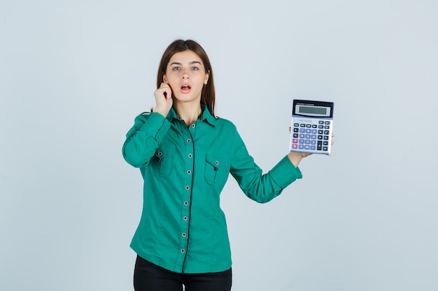 Jovem senhora segurando calculadora enquanto puxa para baixo o lóbulo da orelha em uma camisa verde e parece ansiosa, vista frontal.