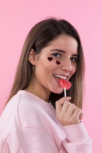 Jovem senhora sorridente com corações de ornamento no rosto e pirulito