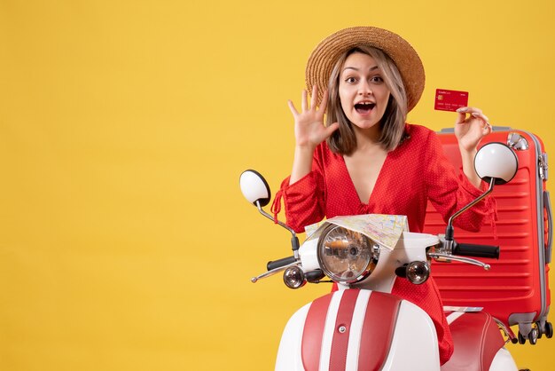 Jovem senhora vestida de vermelho segurando um cartão de crédito e acenando com a mão de frente perto de uma motocicleta