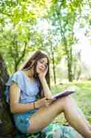 Foto grátis jovem sentada lendo seu livro favorito sobre o green gras sob uma árvore em um belo verão ensolarado