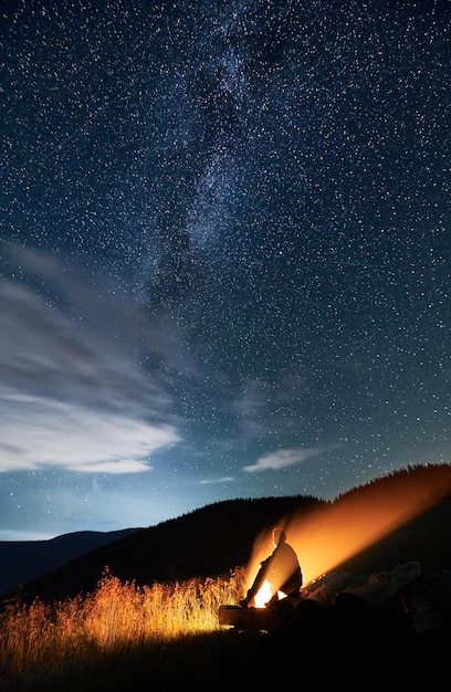 Jovem sentado em toras perto da fogueira nas montanhas sob o céu cheio de estrelas
